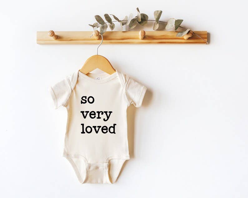 Close-up of “So Very Loved” baby bodysuit in soft natural beige showing the printed message.