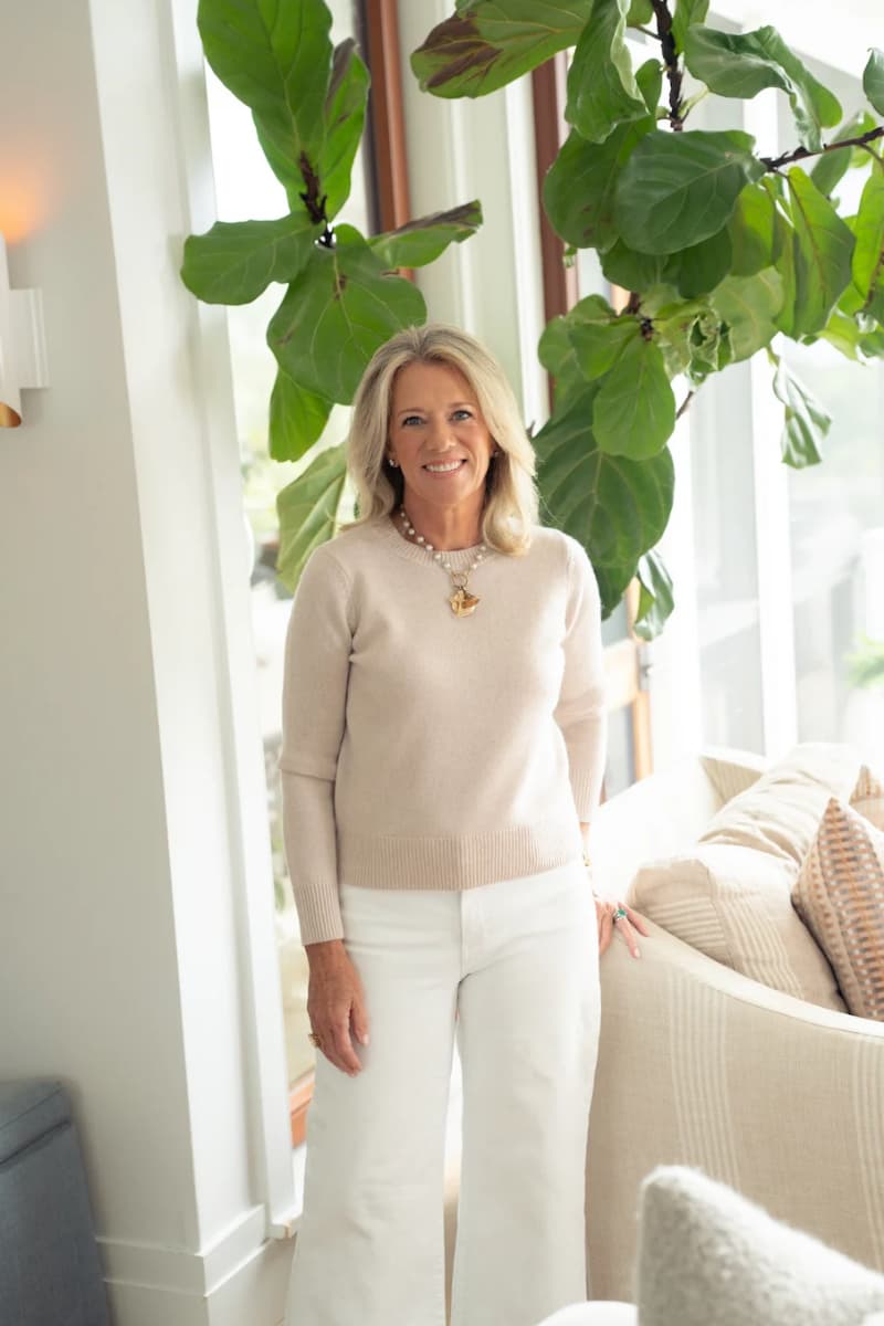 Woman wearing beige crew neck sweater paired with winter white denim and chunky necklace in sundrenched living room.