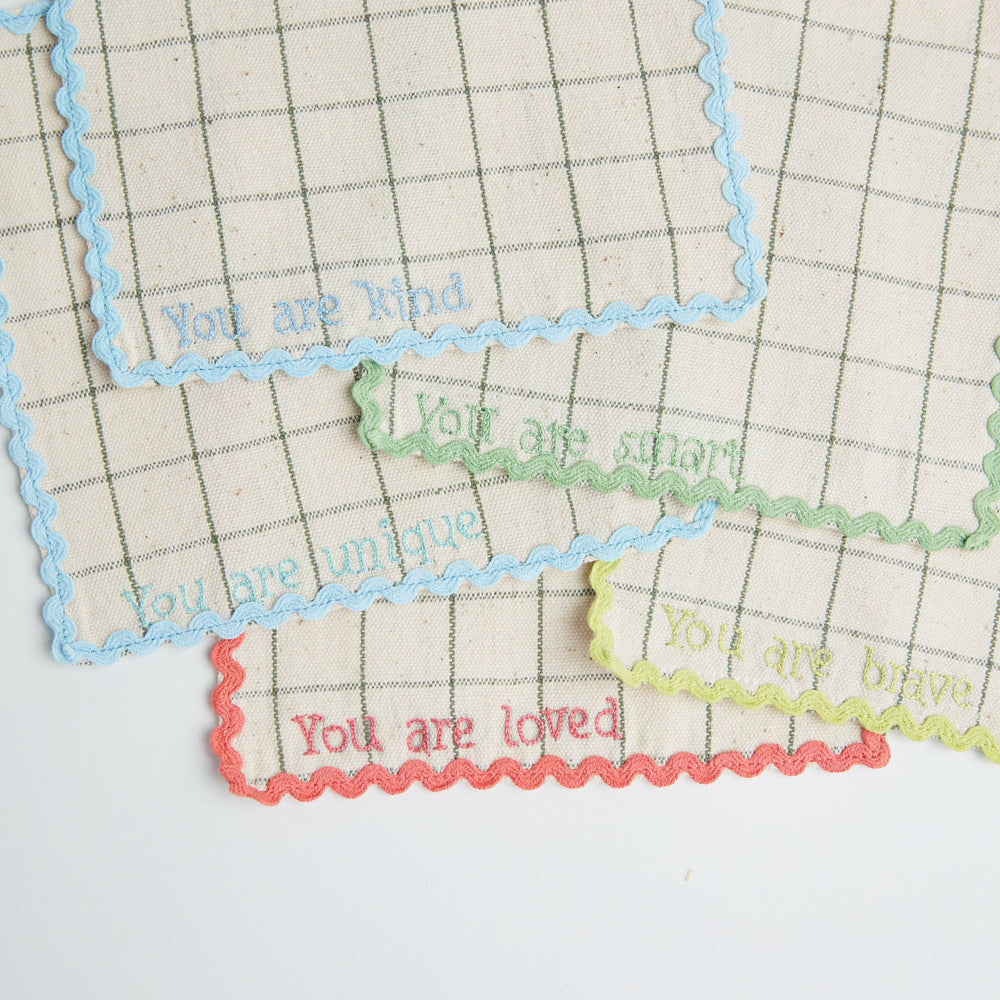 Stack of handwoven lunchbox napkins showing "You Are Loved" embroidery on top napkin in soft, natural tones